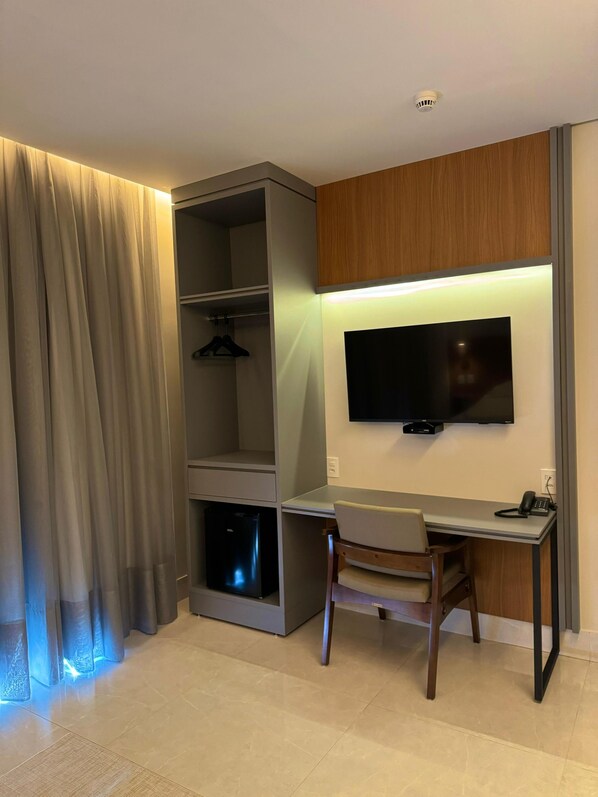 A modern hotel room with a built-in desk, a flat-screen TV mounted on the wall, a wardrobe with a hanging rack, and a compact refrigerator. - Oásis Hotel (Miracema do Tocantins)