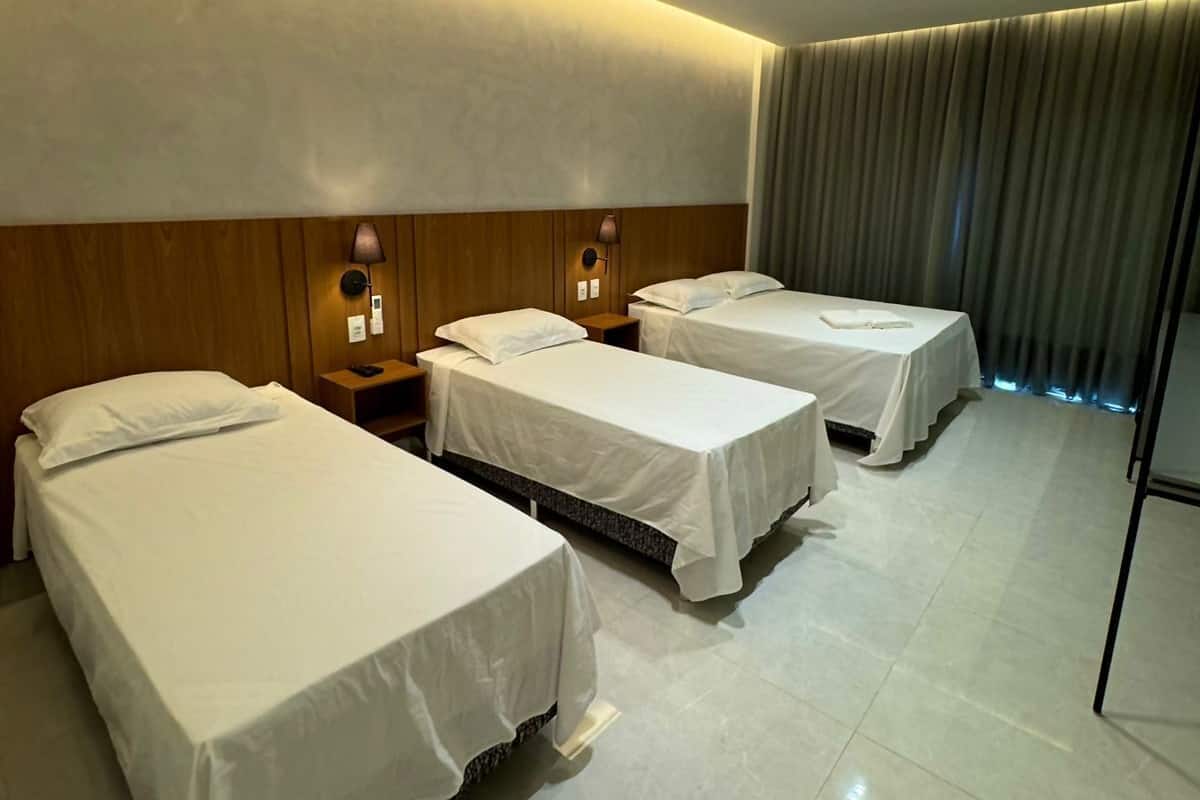 Habitación de hotel con tres camas individuales, cabecero de madera, lámparas de noche y un ventanal con cortinas.