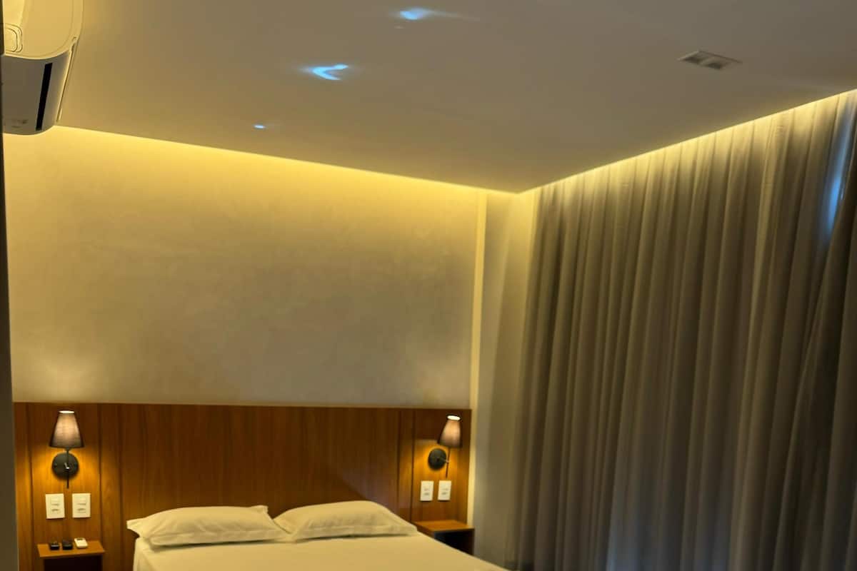 Una habitación de hotel moderna con una cama grande, cabecera de madera, dos lámparas de noche y un ventanal con cortinas.