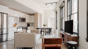A modern kitchen with white cabinets, a microwave, and a stainless steel refrigerator. There is a dining area with a table and chairs, and a sofa in the living room.