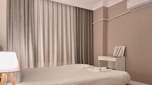 A modern hotel room with a large bed, a bedside table with a lamp, a small bedside table with a picture frame, and a wall-mounted air conditioner.