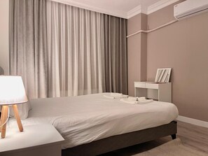 Ett modernt hotellrum med en stor säng, ett sängbord med en lampa, ett mindre sängbord med ett fotoram och en väggmonterad luftkonditioneringsanläggning.