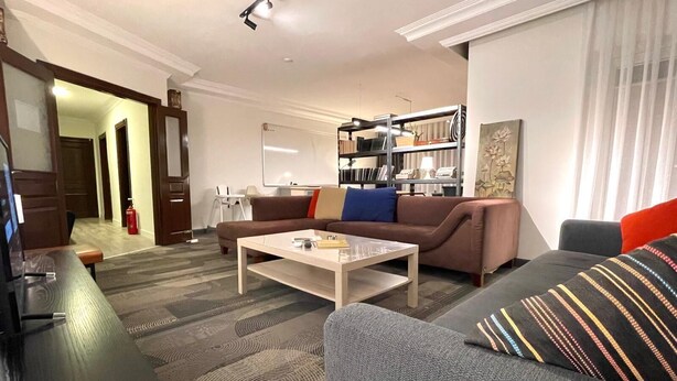 A modern living room with a sofa, a coffee table, and a television. There is a bookshelf and a fireplace in the background.