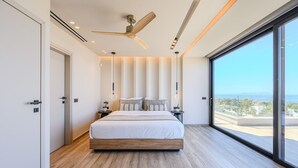 Une chambre moderne avec un grand lit, un ventilateur de plafond et une vue sur la mer grâce à de grandes fenêtres.