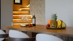 Une table de salle à manger en bois, avec deux verres à vin, une bouteille de vin et une assiette de fruits comprenant de l’ananas, des bananes et du melon.