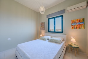 Ein Schlafzimmer mit einem Bett, einem Fenster, einer Wand-Klimaanlage und einem kleinen Tisch mit Lampe und Blumen.