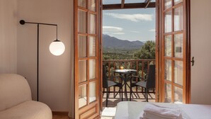 A room with a bed, a round table with chairs, and a balcony with a mountain view.