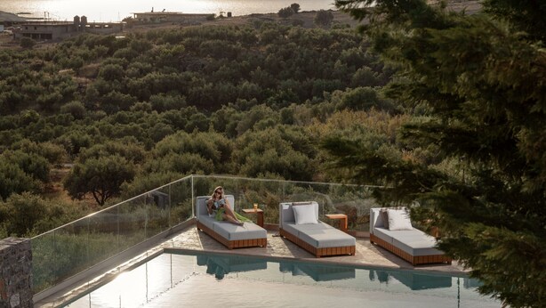 A glass-enclosed infinity pool with two beds and a small table, overlooking a mountainous landscape and the sea.