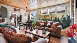 Een gezellige woonkamer met een houten plafond, een bank met dierenkussens, een salontafel en een muurschildering van een bos.