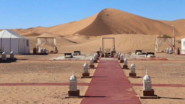 A desert setting with a red carpet leading to a white tent and a dining area with tables and chairs. - Luxury nomadic tented camp  (Merzouga)