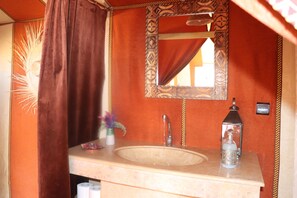 A bathroom with a beige sink, a mirror with a decorative frame, a towel hanging on the wall, and a small vase with a flower. - Luxury nomadic tented camp  (Merzouga)