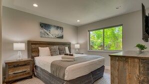 A bedroom with a large bed, wooden nightstands, a painting on the wall, and a window with a view of trees. - NEW! AC, Hot Tub, Grill, Newly Built, Open Floor Plan, Wifi, 6 of 6 – Emerald Bay Way (South Lake Tahoe)