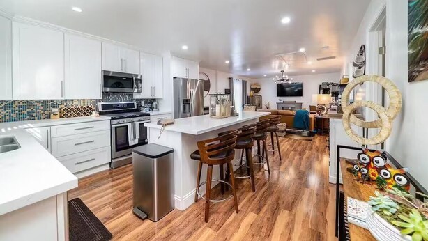 A modern kitchen with white cabinets, stainless steel appliances, a central island with bar stools, and a wooden floor. - Melrose 2 BR w/ large kitchen + fireplace + patio (Vista)