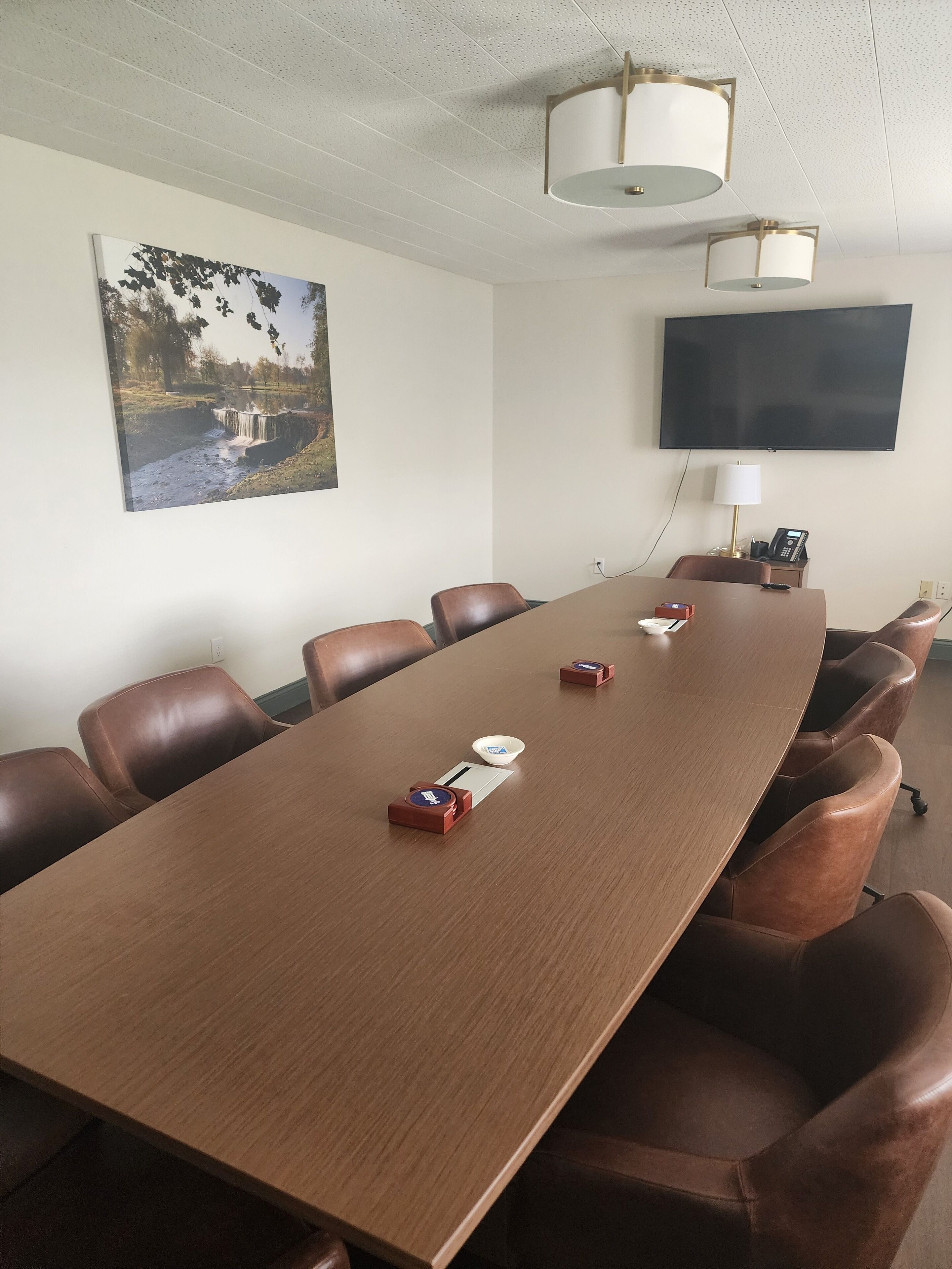A conference room with a long wooden table, brown leather chairs, a mounted TV, and a painting of a bridge over water on the wall.