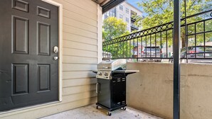 Una parrilla negra sobre ruedas junto a una puerta color beige, con una valla metálica y coches estacionados al fondo.