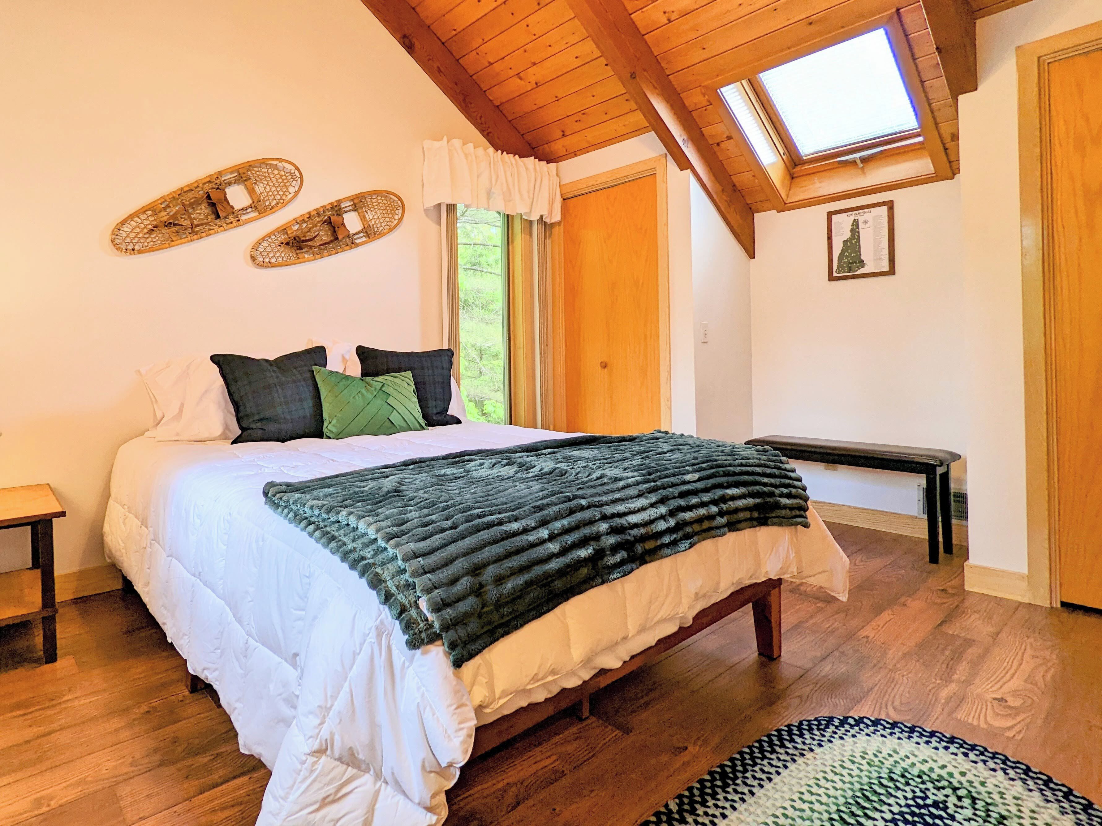 A bedroom with a wooden ceiling, a bed with white bedding and a green throw, a bench, and a skylight.