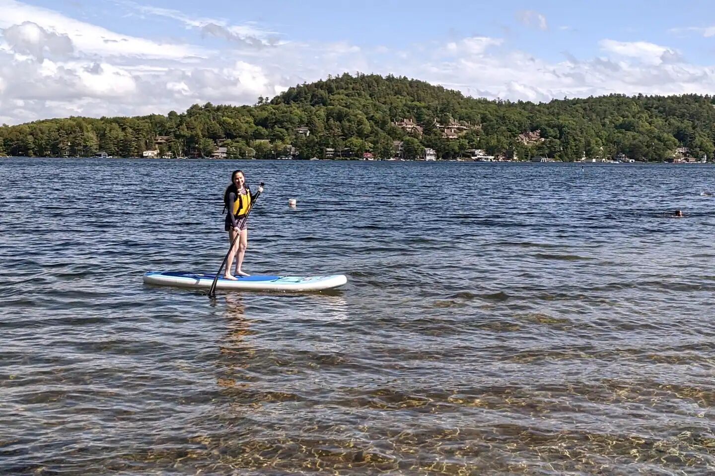 Eine Person steht auf einem Paddleboard auf einem ruhigen See, im Hintergrund ist ein bewaldeter Hügel zu sehen.