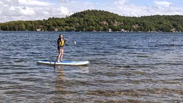 Eine Person steht auf einem Paddleboard auf einem ruhigen See, im Hintergrund ist ein bewaldeter Hügel zu sehen.