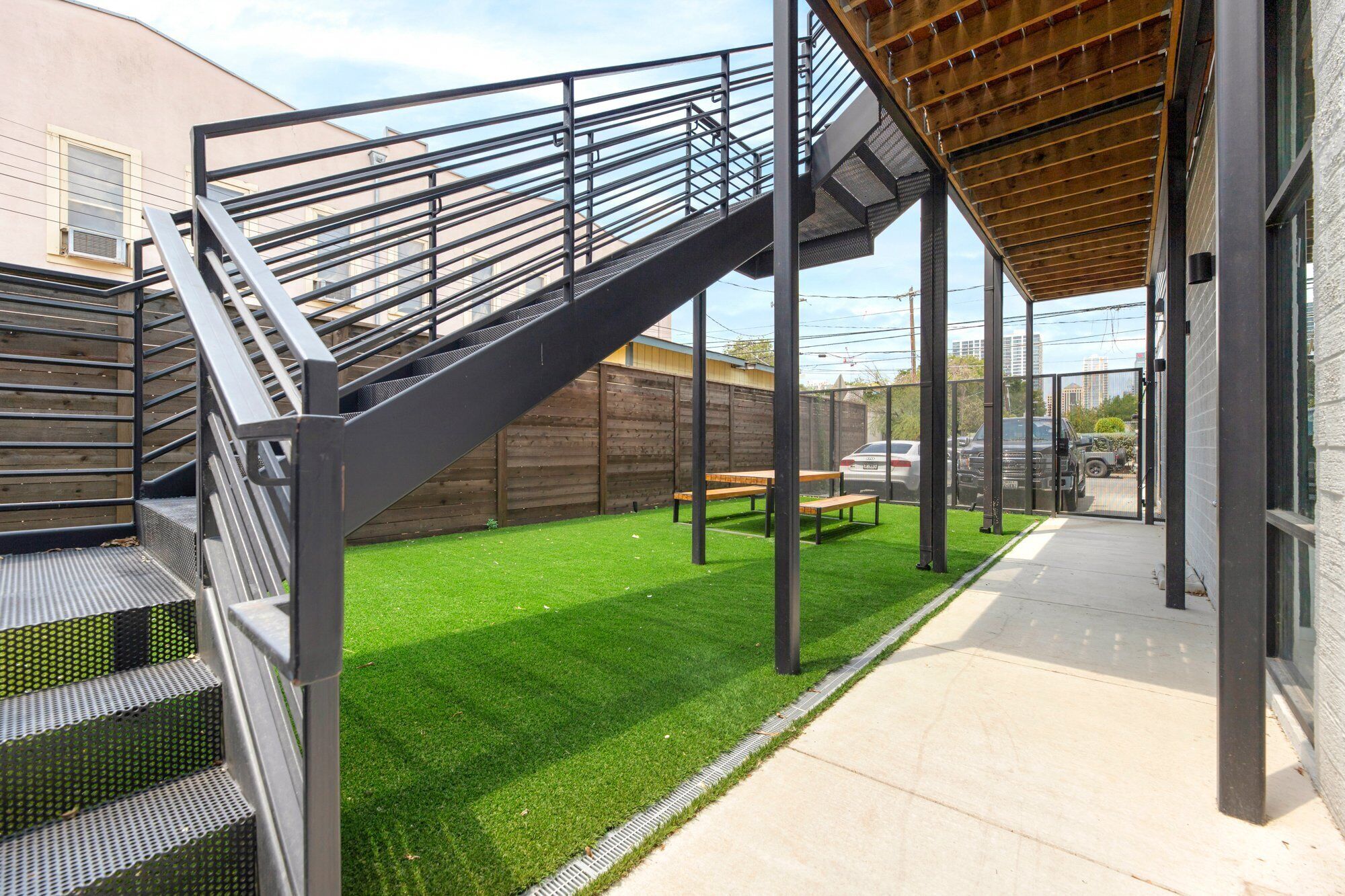 Un espacio exterior cubierto con una escalera metálica que lleva a una terraza de madera, rodeado por un patio cercado con césped artificial y un sendero de concreto.