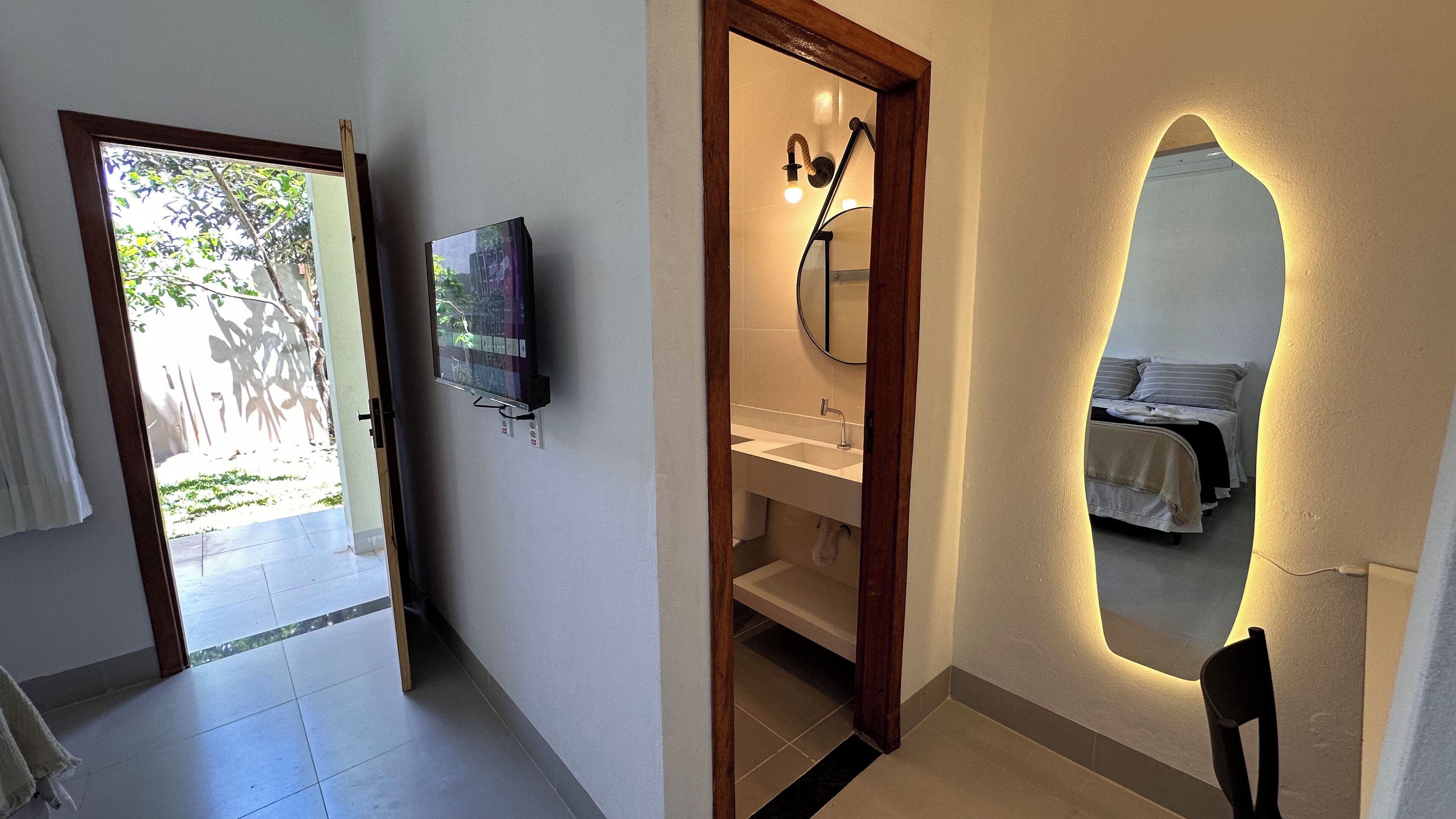 Un bagno con specchio, lavabo e doccia. C'è una TV a parete e una porta che dà su una zona esterna.
