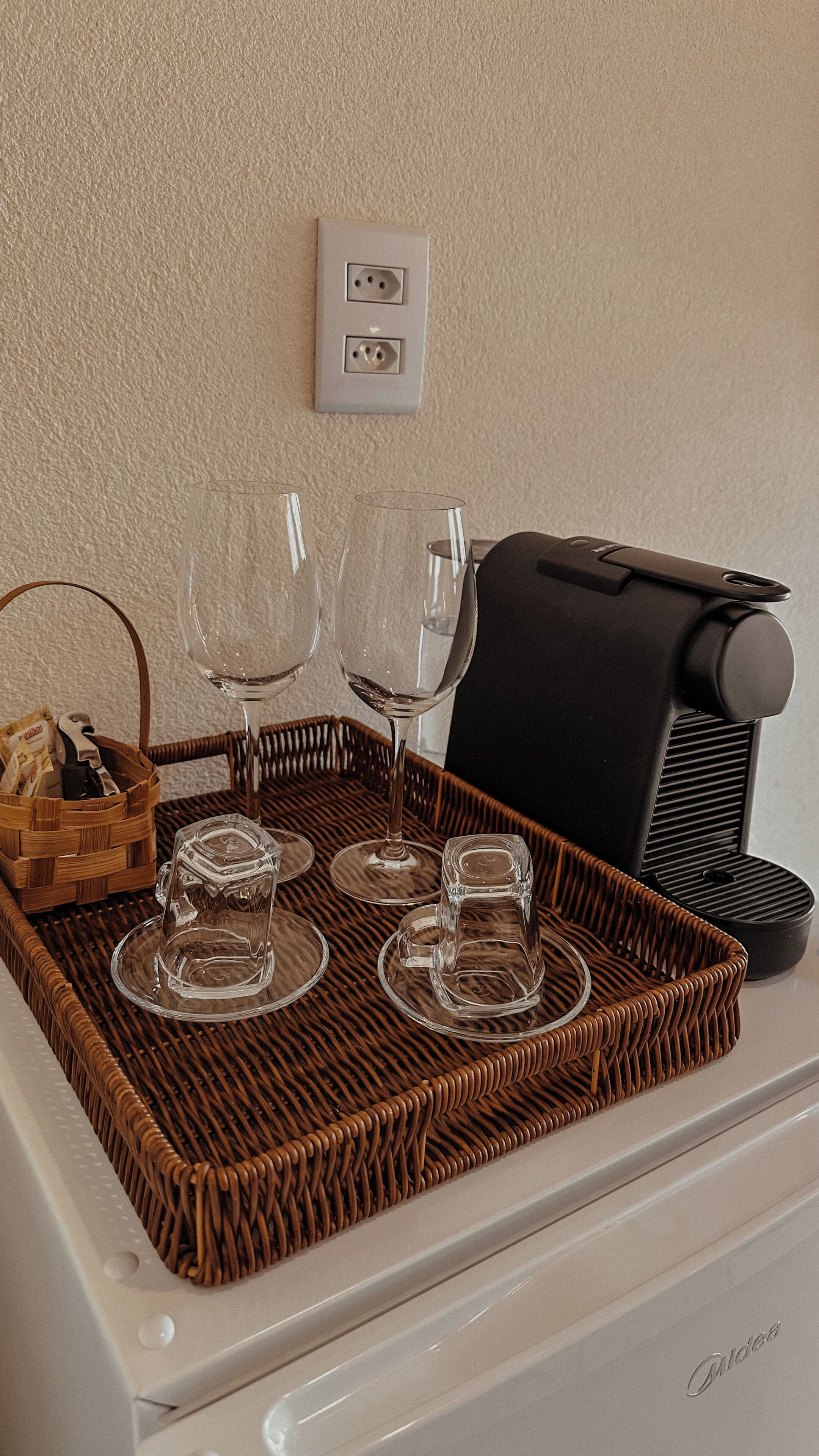 A kitchen area with a coffee maker, a wicker tray with wine glasses, and a basket with utensils.