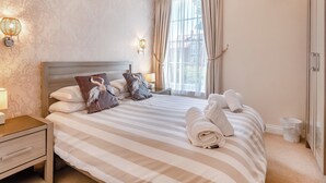 Hotellihuone, jossa on sänky, raidallinen vuodevaatteisto, yöpöytä, lamppu, ikkuna verhoilla ja seinään kiinnitetty valaisin.
