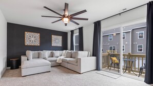 A modern living room with a sectional sofa, ceiling fan, and large sliding glass doors leading to a balcony with outdoor furniture. - Elegant New Home Near Lakes - Relax & Unwind! (Bull Valley)