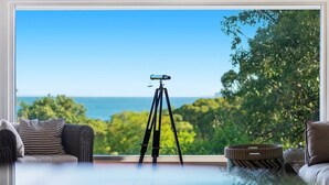 Un télescope, monté sur un trépied, est placé près d’une fenêtre qui offre une vue panoramique sur la mer et la végétation.