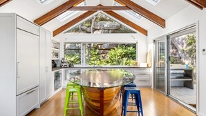 Une cuisine moderne dotée d’un îlot central, de poutres en bois, de grandes fenêtres et offrant une vue sur la verdure.