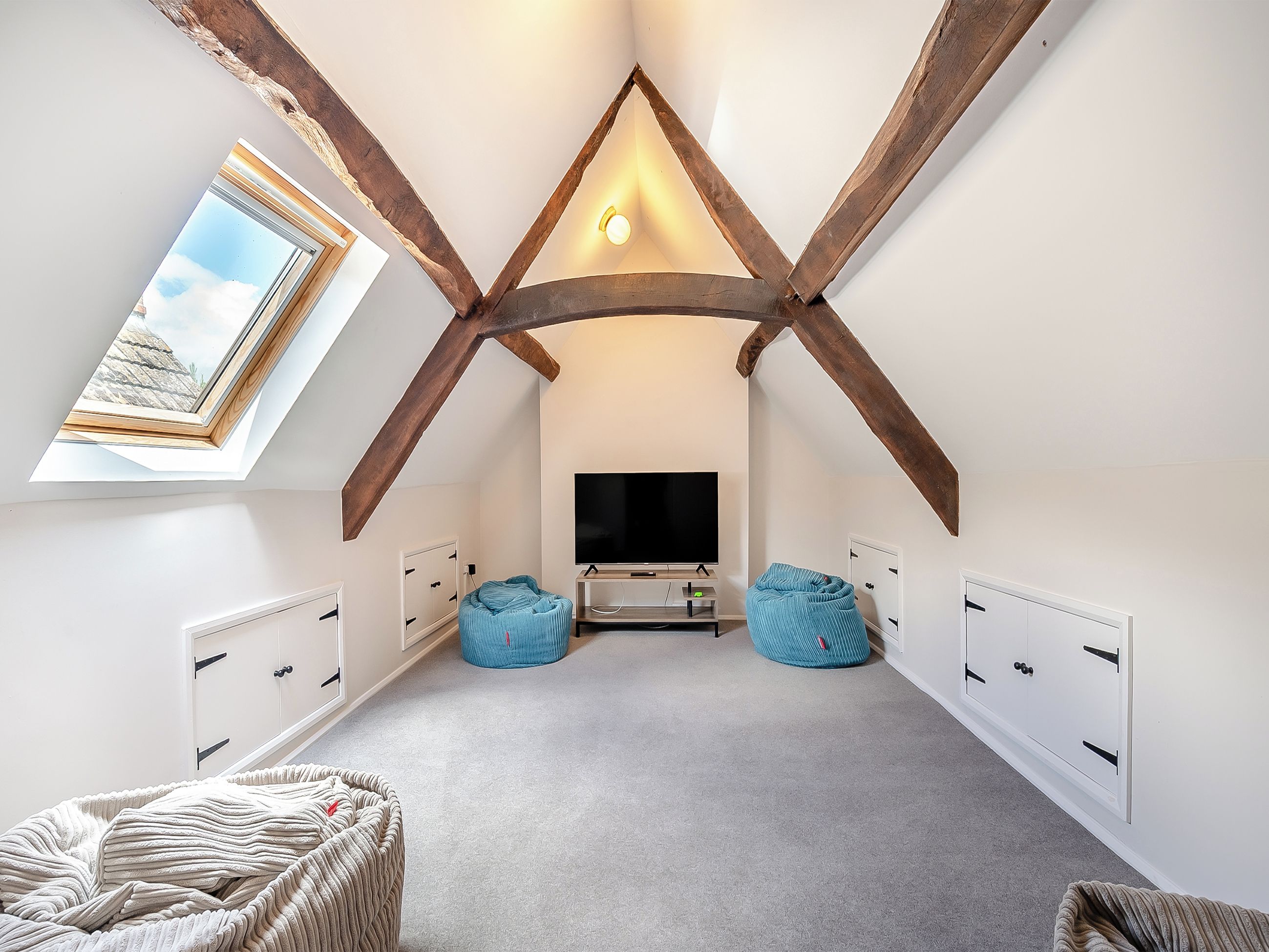 A room with a sloped ceiling, exposed wooden beams, a television, two bean bags, and a window with a view of the sky.