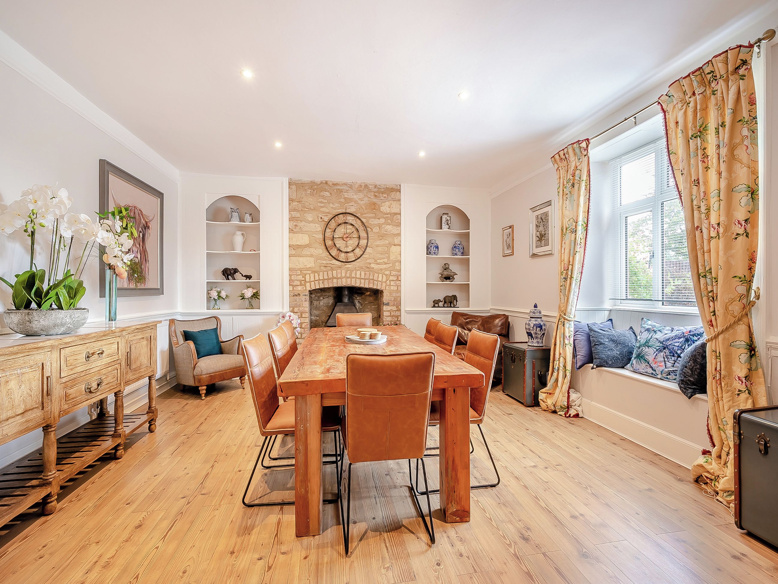 A dining room with a wooden table and chairs, a fireplace, shelves with decorative items, and a window with curtains.