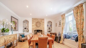 Une salle à manger avec une table et des chaises en bois, une cheminée, des étagères ornées d’objets décoratifs et une fenêtre avec des rideaux.