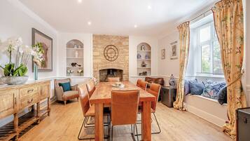 A dining room with a wooden table and chairs, a fireplace, shelves with decorative items, and a window with curtains.
