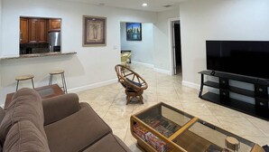 A living room with a sofa, a glass coffee table, a television, and a kitchen area in the background.