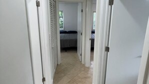 A hallway with white walls and beige tiled flooring leading to a bedroom with a bed and a window.