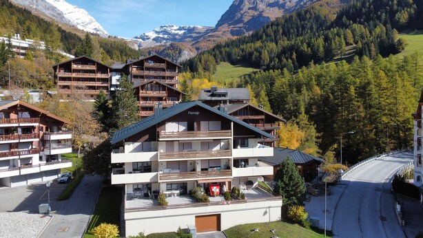 A mountain resort with multiple chalet-style buildings, featuring balconies, a ski lift, and a view of snow-capped mountains.