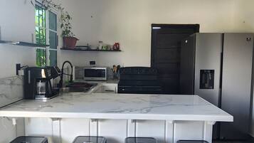 A modern kitchen with a large island, black stools, a microwave, a coffee maker, and a refrigerator.