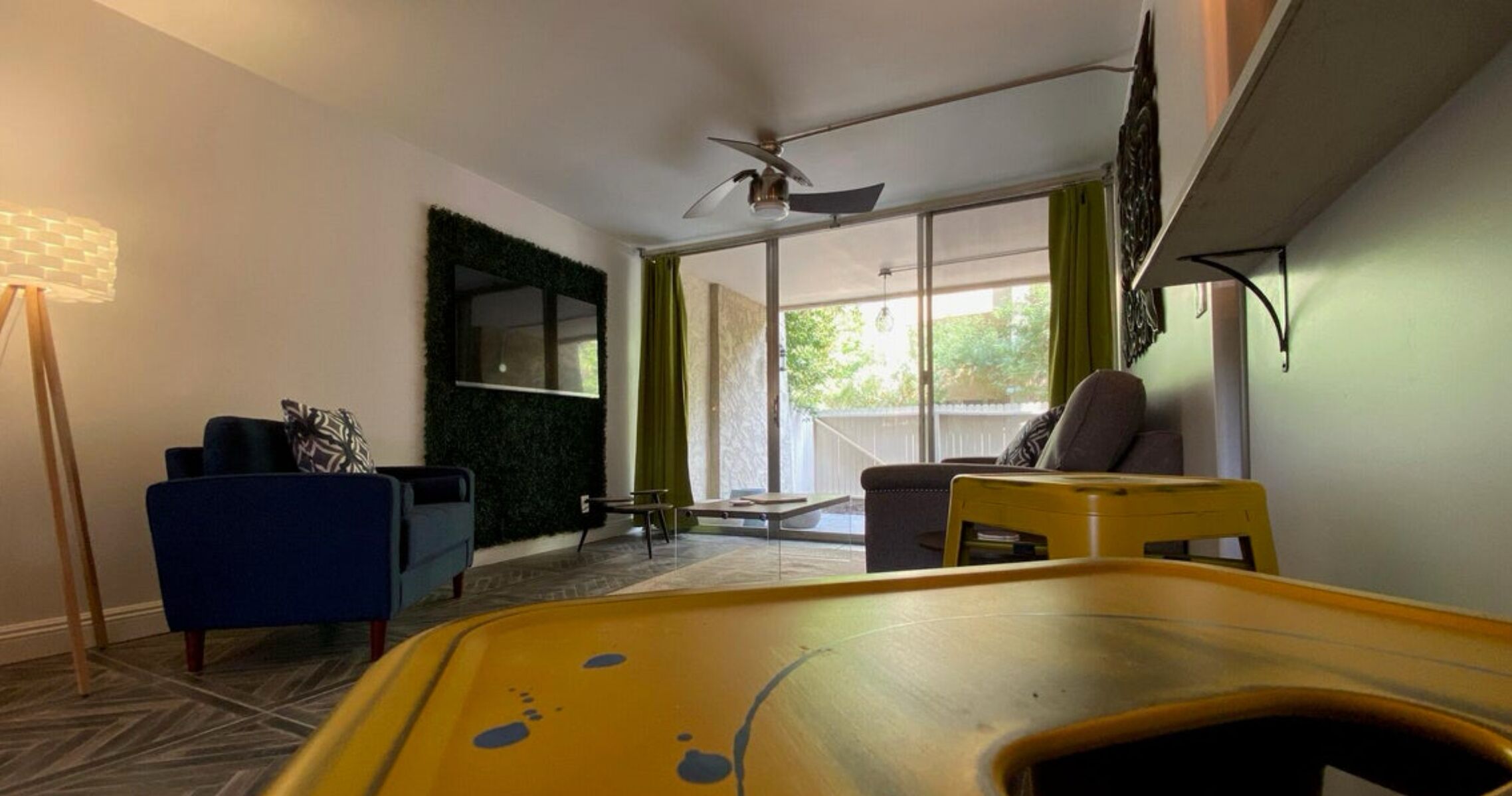 Una sala de estar moderna con ventilador de techo, un sofá azul, una mesa amarilla y una cortina verde.