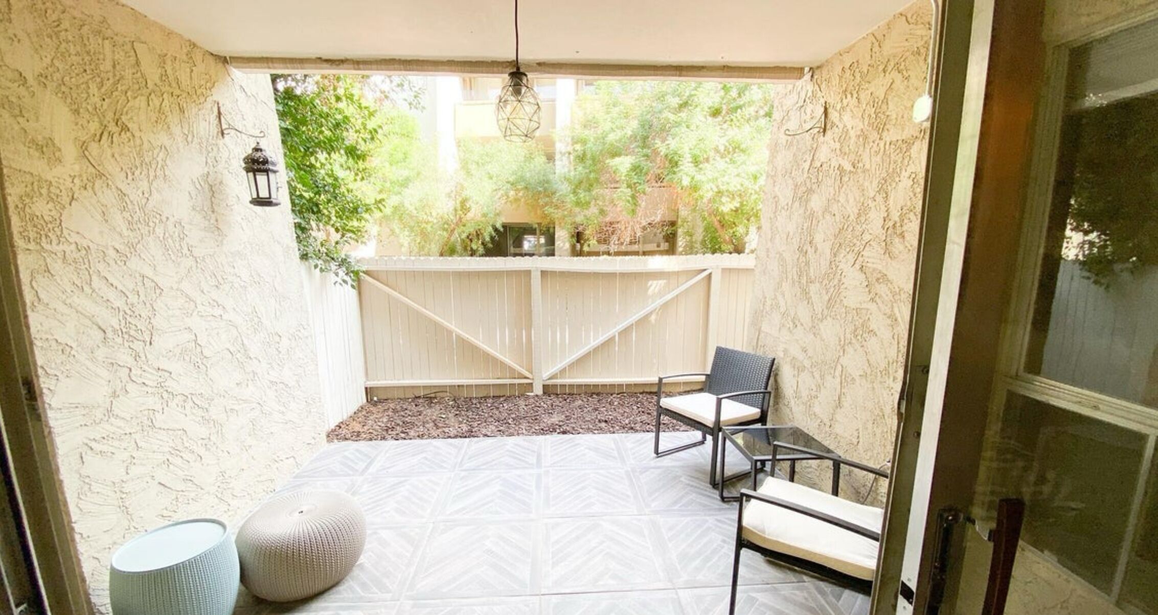 A covered patio with a white patterned floor, two metal chairs, a round cushion, and a lantern.