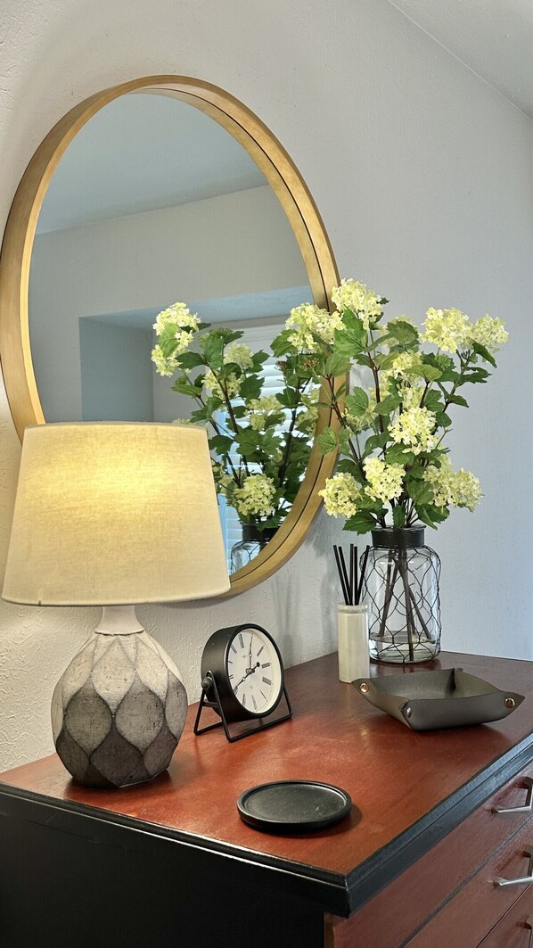 A wooden dresser with a round mirror, a lamp, a clock, and a vase with flowers.