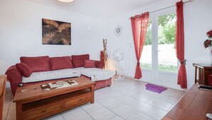 Ein Wohnzimmer mit Ecksofa, Couchtisch aus Holz, Wandventilator und einer Tür mit roten Vorhängen.
