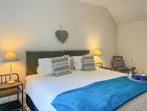 Uma cama bem arrumada com um cobertor azul, duas luminárias de cabeceira, uma decoração de parede em formato de coração e uma pequena mesa com um vaso de flores.