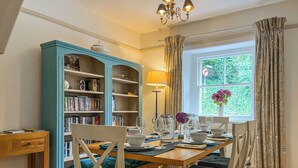 A dining area with a wooden table set for four, surrounded by chairs with cushioned seats. A bookshelf with books and decorative items is to the left. - 3 bedroom accommodation in Bakewell (Bakewell)