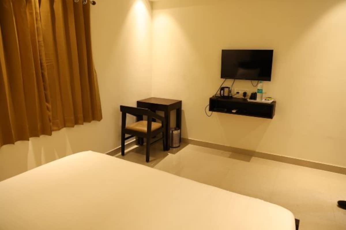 Habitación de hotel con cama, escritorio pequeño, silla, televisión y repisa con objetos.