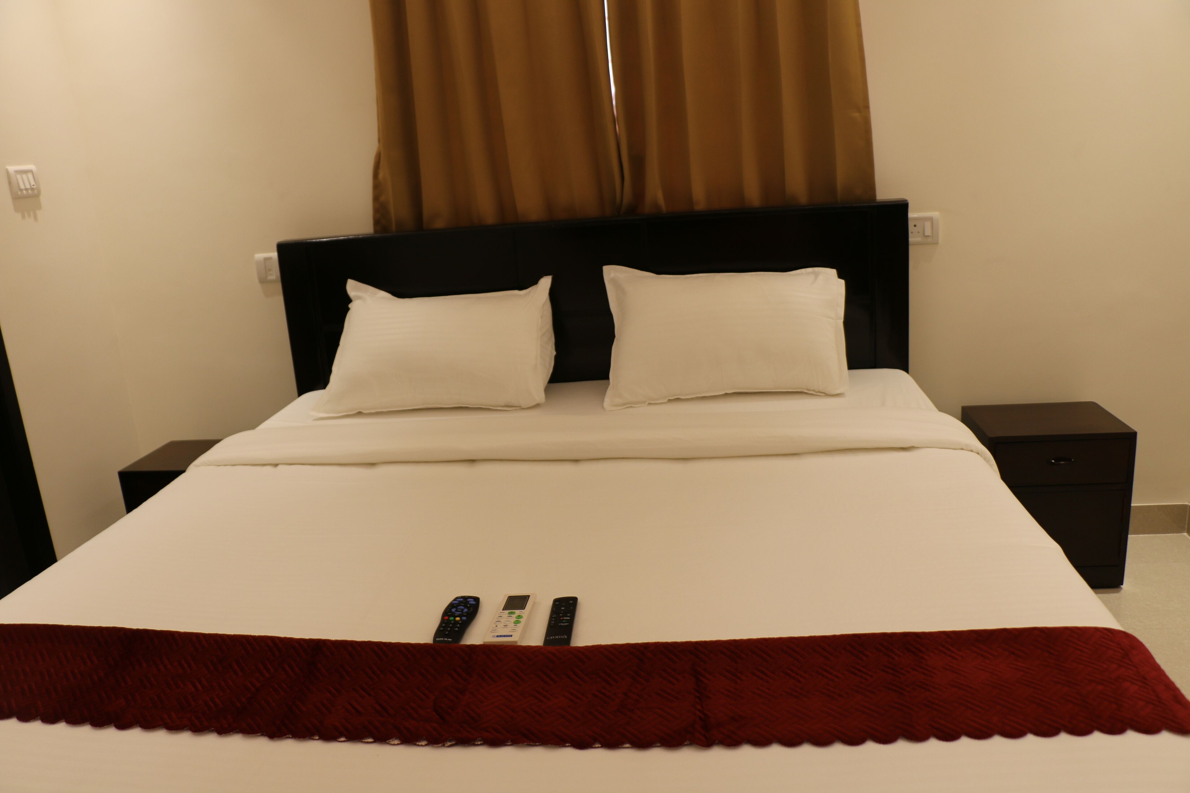 Un lit bien fait, avec du linge de lit blanc et un plaid rouge, deux tables de chevet surmontées de lampes noires, et un rideau marron à la fenêtre.
