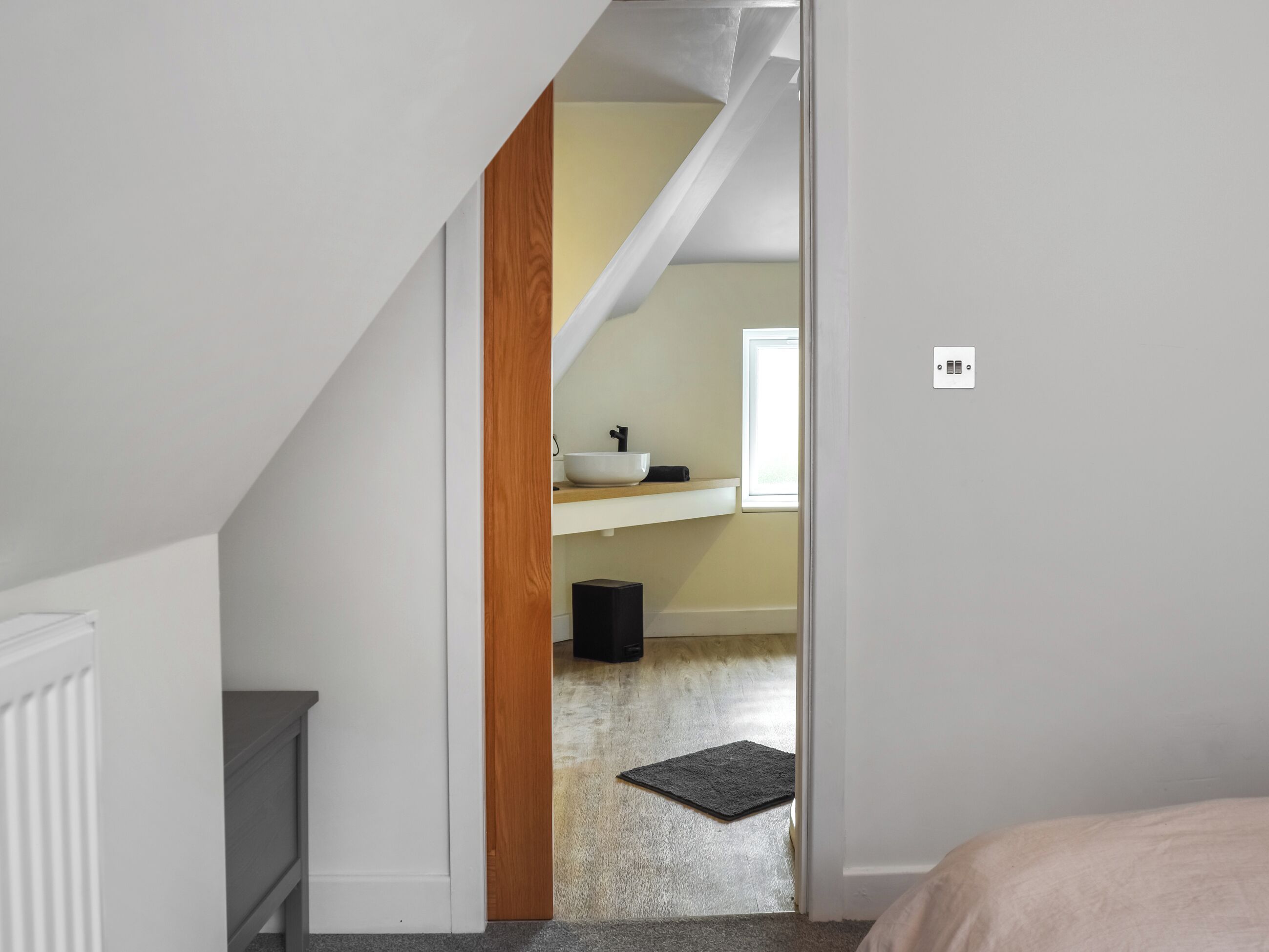 Un dormitorio con cama, puerta que da a un baño con lavamanos y ventana, y una pequeña alfombra en el suelo.