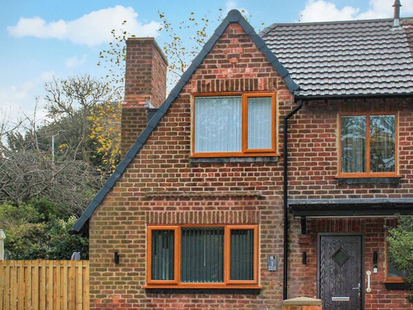 A two-story brick house with a steeply pitched roof, two visible windows with wooden frames, and a chimney.