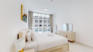 A modern bedroom with a large bed, a bedside table, a dresser, and a window with a city view. - Luxury 1BR Near Miracle Garden |Uni Estate Supreme (Arjan)