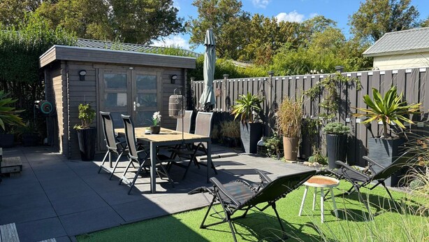 Eine Terrasse mit Tisch und Stühlen, ein kleines Gartenhaus und ein umzäunter Garten.