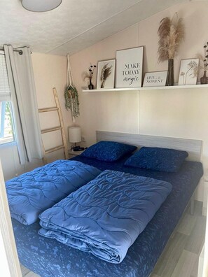 Quarto com cama, mesa de cabeceira, abajur, prateleira com objetos decorativos e janela com cortinas.